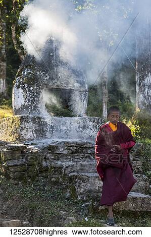 Stock Photograph - A young Buddhist monk outside at Rinchenpong Monastery with a stone altar burning leaves and making a cloud of smoke; Sikkim, India. Fotosearch