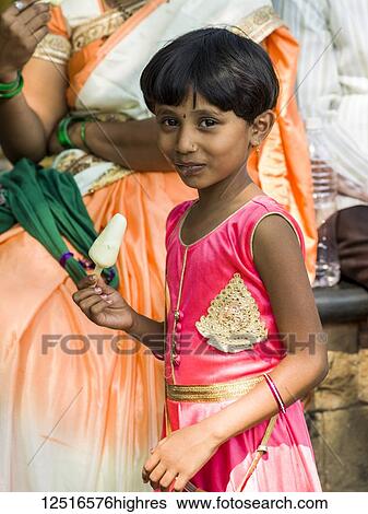Stock Photograph - A young girl in a bright pink dress eating a popsicle and smiling for the camera; Mumbai, Maharashtra, India. Fotosearch