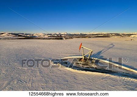 Stock Photograph - Aerial view of pumpjack in a snow-covered field with long shadows, warm light of sunrise, rolling hills, snow-covered mountains and blue sky in the background; Longview, Alberta, Canada. Fotosearch