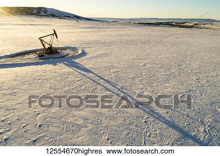 Stock Photograph - Aerial view of pumpjack in a snow-covered field with long shadows, warm light of sunrise, rolling hills, snow-covered mountains and blue sky in the background; Longview, Alberta, Canada. Fotosearch