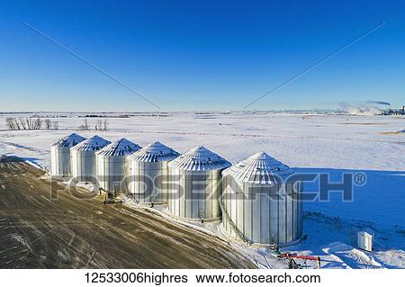 Stock Photograph - Aerial view of snow-covered large metal grain storage bins in a snow-covered field with blue sky, South of Calgary; Alberta, Canada. Fotosearch