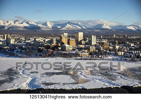 Stock Photograph - Aerial view of snow covering the the sea ice on the frozen shores of downtown Anchorage, the Chugach Mountains in the background beyond the office buildings and hotels in downtown Anchorage in winter, South-central Alaska; Anchorage, Alaska, United.... Fotosearch