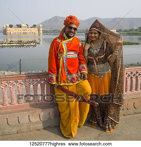 Stock Photograph - An Indian couple stands posing with a grand palace, Jal Mahal, made of red sandstone, submerged in May Sagar Lake in the background; Jaipur, Rajasthan, India. Fotosearch