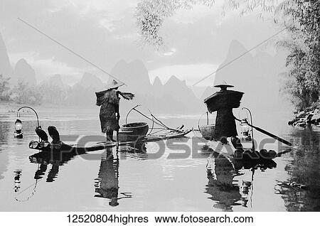 Stock Photograph - Black and white image of fishermen standing on boats on a tranquil river in the fog with karst formations in the distance; China. Fotosearch