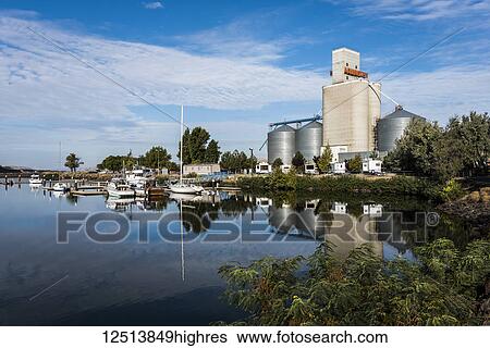 Stock Photograph - Boats mooring along the shoreline of tranquil Lake Umatilla with grain bins on the shore; Arlington, Oregon, United States of America. Fotosearch