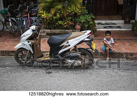 Stock Photograph - Boy sitting along Sisavangvong Road next to a parked motorcycle; Luang Prabang, Luang Prabang, Laos. Fotosearch