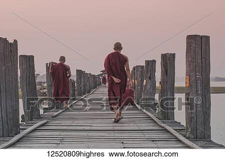 Stock Photograph - Buddhist monks walk down a wooden bridge at dawn; Myanmar. Fotosearch