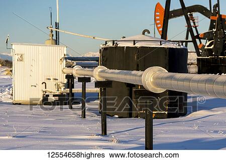 Stock Photograph - Close-up of metal pipeline, with a black metal tank, metal shed and pumpjack in the background in a snow-covered field, West of Airdrie; Alberta, Canada. Fotosearch