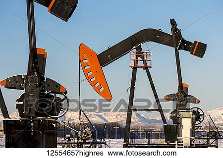 Stock Photograph - Close-up of pumpjack in a snow-covered field with snow-covered mountains and blue sky in the background, West of Airdrie; Alberta, Canada. Fotosearch