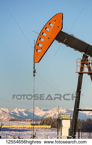Stock Photograph - Close-up of pumpjack with the warm light at sunrise, snow-covered mountains and blue sky in the background, West of Airdrie; Alberta, Canada. Fotosearch