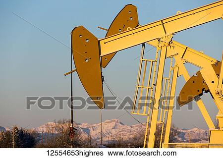 Stock Photograph - Close-up of pumpjacks with the warm light at sunrise, snow-covered mountains and blue sky in the background, West of Airdrie; Alberta, Canada. Fotosearch