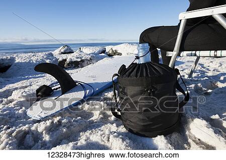 Stock Photograph - Close-up of water bottle and surfboard sitting next to a female surfer on the snowy shore in winter, South-central Alaska; Homer Spit, Alaska, United States of America. Fotosearch