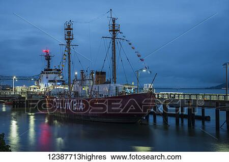Stock Photograph - Columbia Lightship docked along the riverfront with lights illuminating the waterfront at dusk; Astoria, Oregon, United States of America. Fotosearch