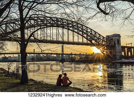 Stock Photograph - Couple Enjoying Sunset Near Hell Gate And Rfk Triboro Bridges, Ralph Demarco Park; Queens, New York, United States Of America. Fotosearch