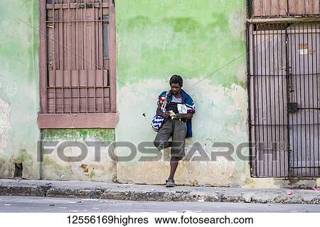 Stock Photograph - Cuban man stands leaning against the wall of a building along a street; Havana, Cuba. Fotosearch