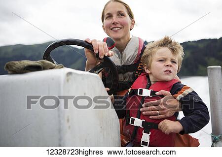 Stock Photograph - Female set-netter driving her skiff while holding her young son, South-central Alaska; Seldovia, Alaska, United States of America. Fotosearch