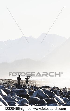 Stock Photograph - Female surfer carries her surfboard over her head along the Homer Spit beach, South-central Alaska; Homer Spit, Alaska, United States of America. Fotosearch