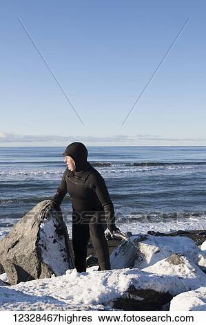 Stock Photograph - Female surfer in a wetsuit walking among the rocks and snow on the Homer Spit shoreline in winter, South-central Alaska; Homer Spit, Alaska, United States of America. Fotosearch