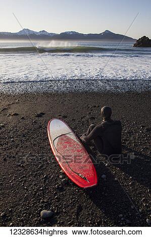 Stock Photograph - Female surfer on the beach looking out at waves and mountains and the Gulf of Alaska, South-central Alaska; Alaska, United States of America. Fotosearch