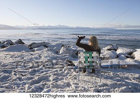 Stock Photograph - Female surfer relaxed in lawn chair on the snowy shore enjoying the view of Kachemak Bay, South-central Alaska; Homer, Alaska, United States of America. Fotosearch