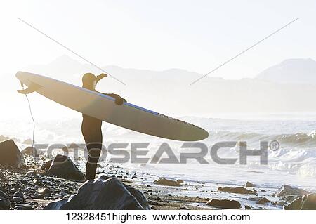 Stock Photograph - Female surfer standing along the wet shore of Kachemak Bay, South-central Alaska; Homer, Alaska, United States of America. Fotosearch