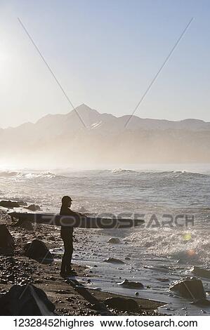 Stock Photograph - Female surfer standing along the wet shore of Kachemak Bay, South-central Alaska; Homer, Alaska, United States of America. Fotosearch