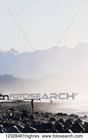 Stock Photograph - Female surfer standing along the wet shore of Kachemak Bay with her surfboard over her head, South-central Alaska; Homer, Alaska, United States of America. Fotosearch