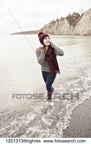 Stock Photograph - Girl standing in shallow water in autumn at the Scarborough Bluffs wearing a knit hat and scarf; Toronto, Ontario, Canada. Fotosearch