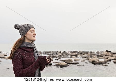 Stock Photograph - Girl standing on a beach in autumn holding a mug and wearing a knit hat and scarf, Woodbine Beach; Toronto, Ontario, Canada. Fotosearch