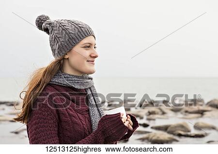 Stock Photograph - Girl standing on a beach in autumn holding a mug and wearing a knit hat and scarf, Woodbine Beach; Toronto, Ontario, Canada. Fotosearch