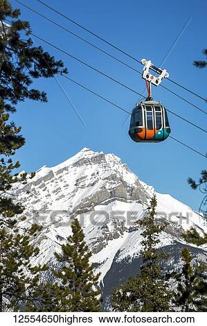 Stock Photograph - Gondola pod travelling along cables with snow covered mountain and blue sky in the background framed by evergreen trees, Banff National Park; Banff, Alberta, Canada. Fotosearch