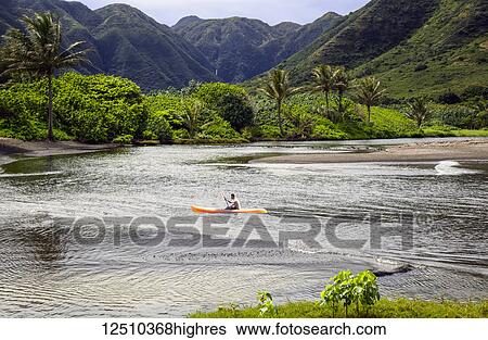Stock Photograph - Kayaker paddles in a bay surrounded by tropical plants and mountains, Halawa Bay, Molokai, Hawaii, United States of America. Fotosearch