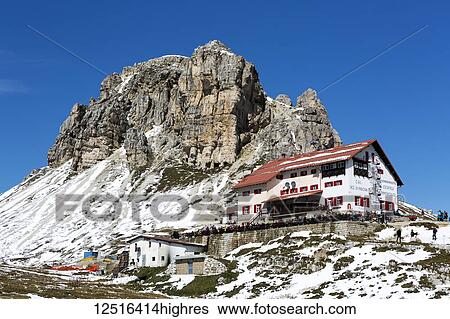 Stock Photograph - Large mountain hotel on a snow-covered rocky ridge with hikers, mountains and blue sky; Sesto, Bolzano, Italy. Fotosearch