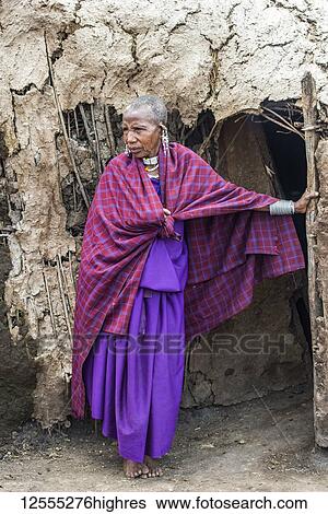 Stock Photograph - Maasai woman elder wearing traditional clothing stands at entrance to her mud hut in the Ngorongoro Conservation Area; Tanzania. Fotosearch