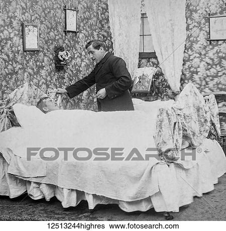 Stock Photograph - Magic lantern slide circa 1900. Victorian. Social History. Image from a slide presentation entitled In His step or In jesus Name. A sick man in his bed wioth the ministertalking to him before he dies. Fotosearch