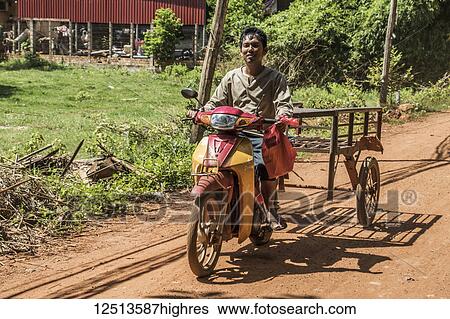 Stock Photograph - Man riding a motorcycle pulling a cart down a dirt road; Siem Reap, Cambodia. Fotosearch