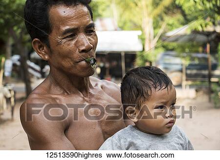 Stock Photograph - Man smoking a cigar holding his daughter in a village near Siem Reap; Siem Reap, Cambodia. Fotosearch