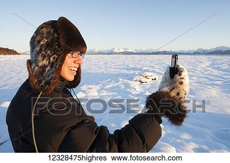 Stock Photograph - Man standing in snow with a fur hat and mittens using a cell phone, South-central Alaska; Alaska, United States of America. Fotosearch