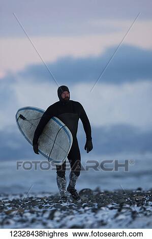 Stock Photograph - Man with surfboard walking on the snow on Homer beach in winter, South-central Alaska, USA. Fotosearch