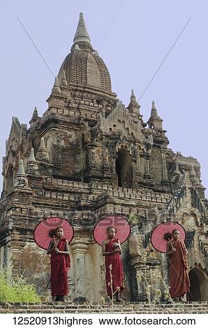 Stock Photograph - Monks with umbrellas stand in front of the ruins of a Buddhist temple; Bagan, Myanmar. Fotosearch
