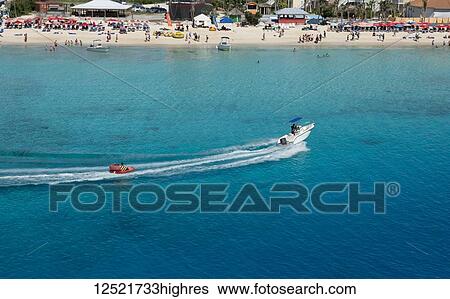 Stock Photograph - Motorboat pulling an inflatable sea through the turquoise waters of the Caribbean Sea in front of a busy beach; Grand Turk Island, Turks and Caicos Islands. Fotosearch