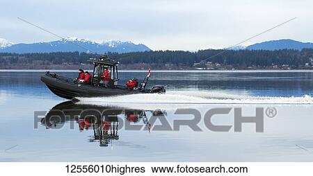 Stock Photograph - Motorboat with four people in red jackets riding along a coastline with the Coast Mountains in the background, Blackie Spit, Crescent Beach; Surrey, British Columbia, Canada. Fotosearch