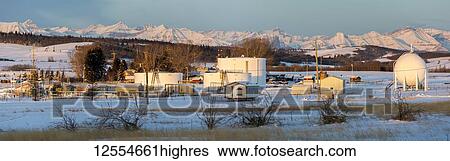 Stock Photograph - Oil storage site with large spherical and round tanks with the warm light of sunrise in a snow-covered field with snow-covered mountains and blue sky in the background, North of Longview; Alberta, Canada. Fotosearch
