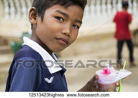 Stock Photograph - Portrait of a boy holding ice shavings and a spoon at Wat Samrong Knong; Battambang, Cambodia. Fotosearch