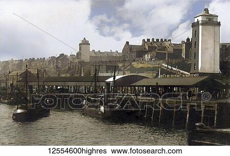 Stock Photograph - Postcard circa 1900, Victorian/Edwardian Social History. A view of North Shields Fish Quay showing steam driven fishing boats, houses and the low and high lights (lighthouses), people walking and working on the fish quay; North Shields, Tyne and Wear,.... Fotosearch