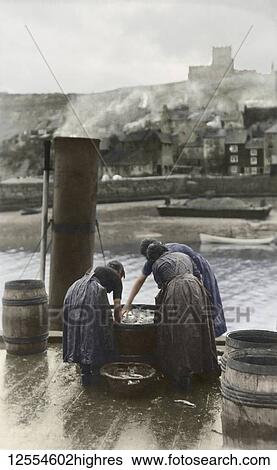 Stock Photograph - Postcard circa 1900, Victorian/Edwardian Social History. Fisherlasses at Whitby unloading and cleaning fish from a steam fishing boat, Whitby church and houses in the background; Whitby,. Fotosearch