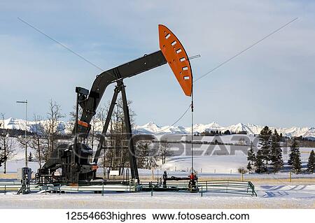 Stock Photograph - Pumpjack in a snow-covered field with snow-covered mountains and blue sky in the background, West of Airdrie; Alberta, Canada. Fotosearch