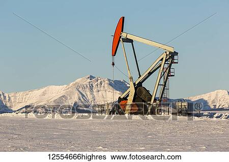 Stock Photograph - Pumpjack in a snow-covered field with snow-covered mountains and blue sky in the background; Longview, Alberta, Canada. Fotosearch