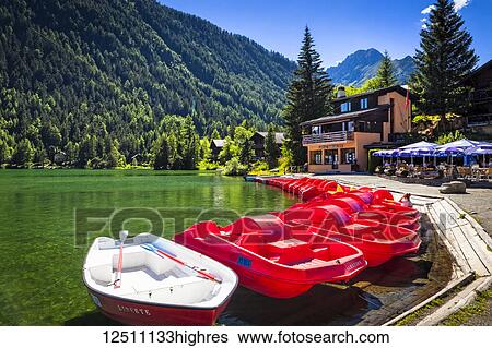 Stock Photograph - Red boats lined up at Champex Lake with an outdoor cafe under blue sky; Champex, Valais, Switzerland. Fotosearch