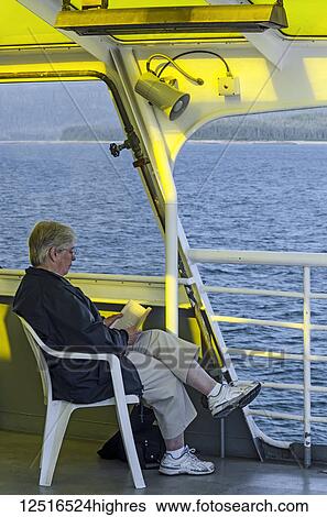Stock Photograph - Senior tourist reading a book in the solarium of the Alaska Marine Highway MV LeConte, Southeast Alaska; Alaska, United States of America. Fotosearch